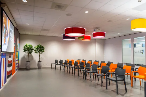 Kleurrijke wachtruimte met zwarte en oranje stoelen, grote ronde plafondlampen en een projectiescherm aan de Niels Bohrweg.